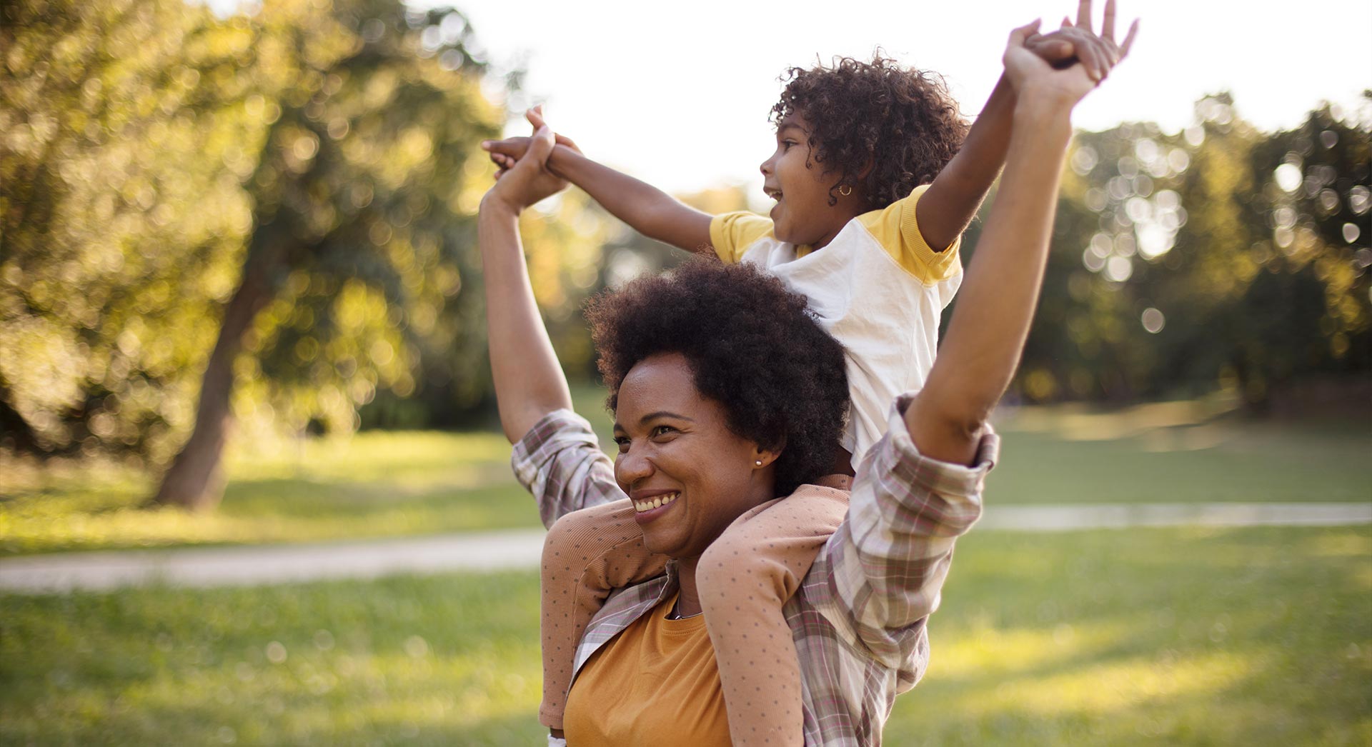 African American mother and daughter standing in park. Little girl on mother shoulders